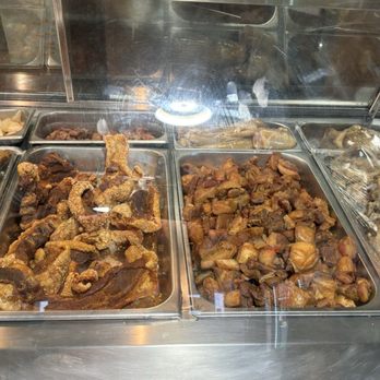 Mr. Meats - Chicharrón - A stainless steel food display case containing different cooked meats and side dishes, including fried pork, stewed pork, and other prepared foods. - Grocery Near Me - Mesa, Arizona