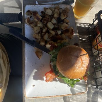 First Watch - A meal is served on a rectangular ceramic plate with roasted cubed potatoes and a burger topped with bacon and fresh greens. A glass of orange juice is placed nearby on a wooden table, with condiments in a wire holder. The setting appears to be outdoors, with sunlight casting shadows on the table. - Breakfast & Brunch Near Me - Mequon, Wisconsin