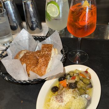 Antico Posto - Aperol + hugo spritz - A table features two cocktails: one with lime slices and a clear liquid, and another with an orange hue and ice. There is a basket of bread lined with parchment paper and a white dish containing an olive salad with black and green olives, grated cheese, and vegetables. The background includes glasses and bar equipment, indicating a restaurant or bar setting. - Italian Near Me - Oak Brook, Illinois