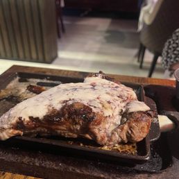 New York Strip - A steak topped with melted blue cheese is presented on a cast iron skillet at a restaurant, with a side of sauce in a small metal cup.