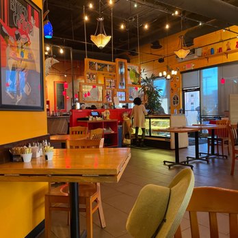 M'tucci's Italian - One of our favorites! - The image shows the interior of a restaurant with warm yellow walls and wooden furniture, including tables and chairs. Decorative hanging lights and artwork adorn the ceiling and walls, creating a vibrant atmosphere. A woman is seen near a display case, possibly selecting food or desserts. Large windows allow natural light into the space, and shelves with colorful vases are visible near the ceiling. The overall setting suggests a lively, inviting dining environment. - Italian Near Me - Albuquerque, New Mexico