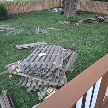 A pile of wooden fencing panels and tools is stacked on the grass in a backyard, with some fencing materials scattered around, suggesting preparations for fencing installation.