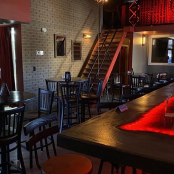 The Overpour - Bar tables and upper wine area - The image shows the interior of a bar with various seating options including round tables and chairs. A staircase leads to an upper level, and a wine display with red lighting is visible above. The brick wall and windows are part of the decor, and the lighting is dim, creating a cozy atmosphere. - Cigar Bars Near Me - El Paso, Texas