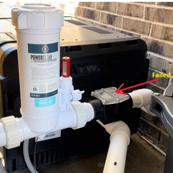 Simonton Pool Consultants - One-way valve, failed or not properly installed. - The image depicts an outdoor water filtration system connected to PVC pipes, situated against a brick wall. A filter unit labeled 'PowerClean' is mounted on the system. A black component with a transparent cover is attached to the piping, with a red label indicating a failed one-way valve, marked with yellow text. The setup appears to be part of a water treatment or pool system, with additional equipment visible nearby. - Pool & Hot Tub Service Near Me - Sealy, Texas