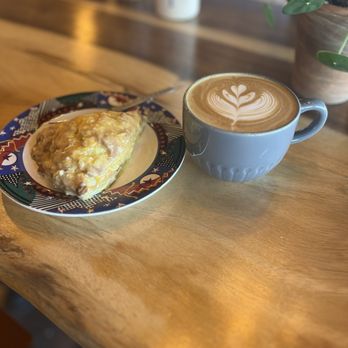 Ascend Coffee - A latte with foam art and a pastry on a decorative plate are placed on a wooden table in a coffee shop setting. - Coffee & Tea Near Me - Indianapolis, Indiana