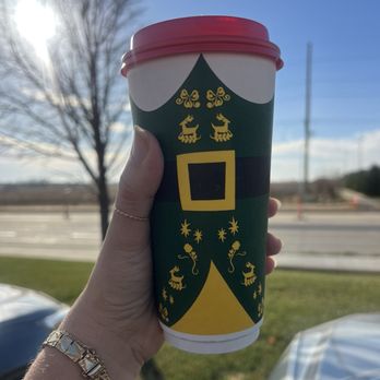 Scooter's Coffee - Large cappuccino - A hand is holding a large disposable coffee cup decorated with a Christmas elf costume design, featuring green, yellow, and black colors. The background shows a clear blue sky, a leafless tree, and a road with some traffic cones, indicating an outdoor setting on a sunny day. The person is wearing a bracelet and a ring, and the scene suggests a casual moment during winter or the holiday season. - Coffee & Tea Near Me - Cedar Falls, Iowa