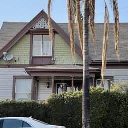 Before - A two-story house features a gabled roof with shingles, siding in light colors, and a small front porch supported by columns. The house has multiple windows, including a prominent upper window with curtains. A palm tree stands in front, with some dried fronds hanging down. The property is bordered by bushes and a hedge, with a partial view of a parked car in the foreground. A satellite dish is mounted on the house exterior.