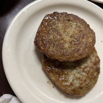 Red Mountain Cafe - Two sausage patties are arranged on a plain white plate, cooked to a golden brown with a slightly crispy exterior. The patties appear juicy and well-cooked, with some visible fat and seasoning. The plate is on a dark surface, with part of a napkin visible in the corner. The image focuses on the sausage patties, which are a common breakfast item. - American Near Me - Mesa, Arizona