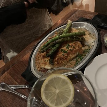 Public Kitchen - Baked haddock - The image shows a metal platter with baked haddock garnished with chopped herbs and roasted asparagus on top. The dish is placed on a wooden table in a restaurant setting. In the foreground, there is a glass of water with a lemon slice. Part of a white plate and a small bowl are also visible. The background includes a person wearing a knitted sweater and other restaurant furniture. - Gastropubs Near Me - Wakefield, Massachusetts