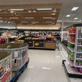 Target - The image shows an interior view of a grocery store aisle with produce sections in the foreground, including watermelons in wooden bins. To the right, there are snack shelves stocked with various chips and snack items, with a visible price sign. The background includes more grocery shelves, a ceiling with wooden panel accents, and a few shoppers shopping in the store. The floor is shiny and clean, reflecting the store's lighting. - Department Stores Near Me - Arlington Heights, Illinois