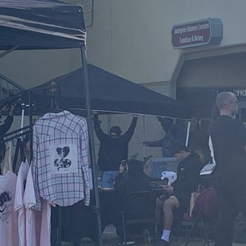 Discovery Records - Multiple individuals are gathered outside a record store, with some standing under a canopy and others seated. Merchandise such as T-shirts and a shirt with a graphic are displayed on a stand. The store's signage indicates it offers entertainment, music, and related services. The scene appears to be casual, with people engaging in various activities, including taking photos and browsing. - Vinyl Records Near Me - Temecula, California