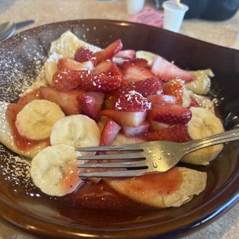 Weck's - Strawberry crepes - A dark, oval-shaped bowl contains strawberry crepes topped with sliced bananas, fresh strawberries, and a dusting of powdered sugar. The crepes are folded and appear soft, with some strawberry syrup visible. The setting includes a spoon and fork on a beige table, with some cups and glasses in the background, indicating a restaurant environment. - Breakfast & Brunch Near Me - Santa Fe, New Mexico