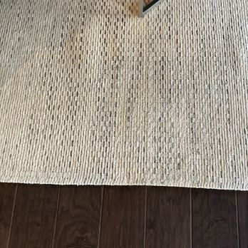 I Clean Carpets - Stain is completely gone. - The photograph depicts a beige, textured rug with a pattern of small, elongated shapes, placed on a dark brown wooden floor. The rug's edge is visible in the upper right corner, and part of a metal frame or furniture leg is also seen. The wooden floor has a smooth finish with visible wood grain and plank lines, contrasting with the rug's textured surface. - Carpet Cleaning Near Me