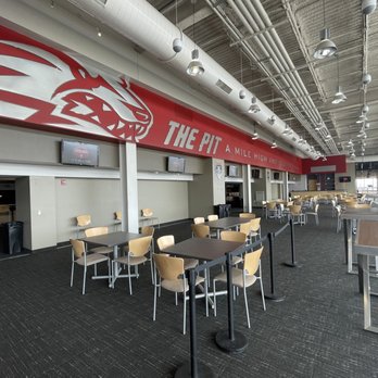 The Pit - Club Level @ the Pit! - The image depicts an interior seating area inside The Pit sports arena, featuring several tables and chairs arranged in the space. The area has a high ceiling with exposed ductwork and lighting fixtures. A prominent red sign with a logo and the text "THE PIT" is visible on the wall, along with additional signage. The space appears to be a lounge or club level area, with large windows on one side allowing natural light to enter. - Stadiums & Arenas Near Me - Albuquerque, New Mexico