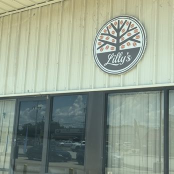 Lilly’s - Outdoors - The photo captures the front exterior of Lilly's, a coffee shop, with a metal siding building and a circular logo sign displaying a stylized tree with coffee beans as leaves and the name Lilly's written in cursive. Large glass windows reflect the parking lot and street scene outside, including cars and power lines. The building has a metal roof with a light fixture visible in the upper left corner. - Coffee & Tea Near Me - Willis, Texas