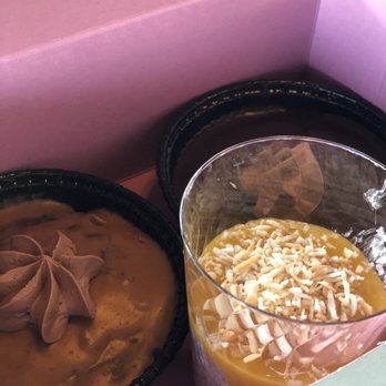 Sweetspot - Peanut Butter Brownie,Chocolate Brownie, and custard. - The image shows two small black plastic dessert cups and a layered dessert in a clear glass container, all placed inside a pink box. The layered dessert appears to have a yellow topping with white shavings, possibly white chocolate, and a creamy base. The setting suggests these are desserts prepared for takeout or delivery, with the pink box serving as packaging. - Cupcakes Near Me - Redding, California