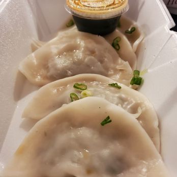 Somac - Steamed beef dumplings - A foam takeout container holds three steamed beef dumplings topped with chopped green onions. A small cup of soy sauce is included for dipping. The dumplings have a smooth, slightly shiny surface, indicating they are freshly steamed. The container appears to be set on a dark surface, possibly outdoors or in a casual dining setting. - Chicken Wings Near Me - Chantilly, Virginia
