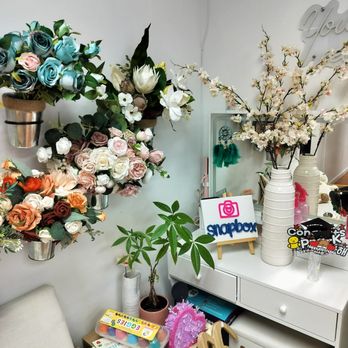 Snap Box - The props - The image shows a corner with a white dresser holding decorative items including artificial flowers in vases, a small potted plant, and a Snapbox sign. There are additional items like books, a pink decorative object, and a pair of shoes on the floor. The background features a plain white wall with some wall decor and a chair partially visible. - Session Photography Near Me - Flushing, New York