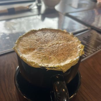 Masuda’s Cafe - Pistachio cardamom coffee - A black ceramic mug containing pistachio cardamom coffee with a foamy surface and a dusting of spices or ground nuts. The mug is placed on a matching black saucer on a wooden table, with a window in the background allowing natural light to illuminate the scene. The setting appears to be inside a cafe, with some blurred objects and a plant visible near the window. - Cafes Near Me - Amherst, Massachusetts