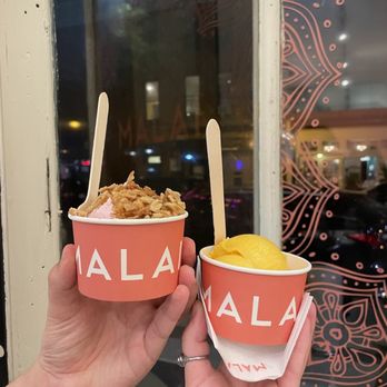Malai - Rose and Apricot Mace - Two cups of ice cream from Malai in Brooklyn, NY, are held by hands near a window with decorative patterns. One cup has a pink ice cream with toppings, and the other has yellow ice cream. - Ice Cream & Frozen Yogurt Near Me - Brooklyn, New York
