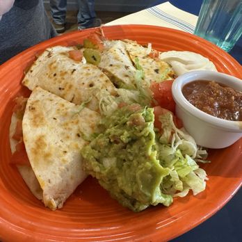 Pantry Restaurant - Quesadilla - A large orange plate contains quesadillas made of folded tortillas with visible fillings, accompanied by a serving of guacamole, a small bowl of salsa, and a dollop of sour cream. The setting appears to be a restaurant with a dark table, a glass of water, and utensils nearby. The food presentation suggests Mexican cuisine, featuring traditional ingredients like guacamole and salsa. - Breakfast & Brunch Near Me - Santa Fe, New Mexico