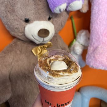 Mr Bear - Jasmine Dream Milk Tea - A caramel milk tea with whipped cream and cinnamon topping is held in front of plush teddy bears in a store display. - Desserts Near Me - Queens, New York
