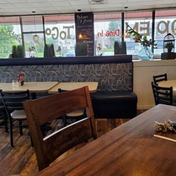 Inside - A restaurant interior featuring tables, chairs, a booth with patterned upholstery, and decorative plants near large windows with signage.