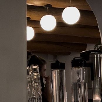 La Reina - Bartender with no mask and eating behind the bar while on shift and was rude. Touching her nose. Not washing hands afterwards. - A person is partially visible behind a bar counter, touching their nose, with glassware and beverage dispensers in the foreground. The background features hanging spherical lights and a wooden ceiling. - Bars Near Me - Santa Fe, New Mexico