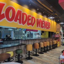 the inside of a fast food restaurant - Inside Loaded Wiener, a fast-casual hotdog restaurant, there is a counter with multiple colorful stools in shades of yellow, green, and brown. Behind the counter, the kitchen area is visible with staff preparing food. A large yellow sign with red lettering spelling 'LOADED WIENER' is prominently displayed above the counter. The interior features wood flooring and modern lighting fixtures, creating a lively atmosphere.