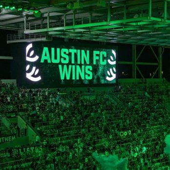 Q2 Stadium - The image features a large digital screen at Q2 Stadium, illuminated with the message 'AUSTIN FC WINS' in bright green text. The stadium is filled with spectators seated in the stands, and the overall scene is bathed in green lighting, indicating a celebratory moment after a soccer match. - Stadiums & Arenas Near Me - Austin, Texas