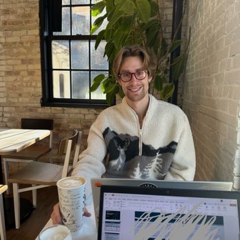 Jones Coffee - A man with glasses, wavy hair, and a light-colored sweater with a black and white animal pattern is sitting at a table in a coffee shop. He is smiling at the camera, with two drinks on the table, one in a paper cup and the other in a glass. The background includes a large window with black framing, a brick wall, and a tall leafy plant. The setting appears to be a modern, industrial-style coffee shop with natural light. - Coffee & Tea Near Me - Chaska, Minnesota