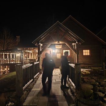 Johnny Seesaw's - Two individuals are standing on a stone pathway leading to a log cabin at night, with warm lighting illuminating the entrance. - American Near Me - Peru, Vermont