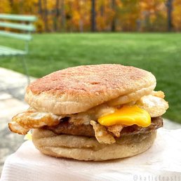 Breakfast Sandwich - A breakfast sandwich featuring a fried egg, crispy bacon, and melted cheese on an English muffin, set outdoors with a grassy background and autumn trees.