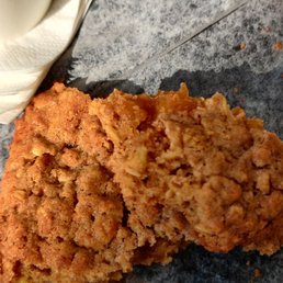 Oatmeal Cookie - A partially eaten apple oatmeal cookie with a crumbly texture rests on parchment paper. The cookie appears golden brown and moist inside. The setting includes a wooden table, with a white mug and napkins partially visible in the background, suggesting a casual dining or coffee shop environment.