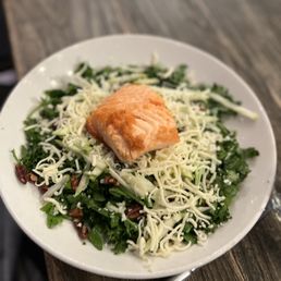 Kale & Apple Salad - A salad featuring leafy greens, shredded cheese, a piece of cooked salmon, and chopped nuts on a white plate, served on a wooden table.