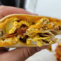 Breakfast Burrito - A photograph shows a breakfast burrito with a filling of scrambled eggs, sausage, and melted cheese, partially unwrapped and held in a person's hand. The background includes a blurred view of a food container and a piece of fried food. The image emphasizes the cross-section of the burrito, highlighting the ingredients inside.