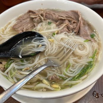 Devoted - Beef pho. Huge portion. - A large white bowl contains beef pho with rice noodles, thinly sliced beef, bean sprouts, chopped green onions, and microgreens in a clear broth. A black spoon and a fork are placed in the bowl, and a person's hand is visible in the background. The bowl is on a wooden table, with a glass bowl and condiments nearby. - Ramen Near Me - Stillwater, Oklahoma