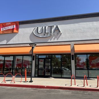 Davis Collection - The image shows the exterior of an Ulta Beauty store with a modern facade. The storefront features large glass windows, orange awnings above the entrance, and a prominent sign with the word "ULTA" in metallic letters. A red banner indicating "opening soon" is attached near the roof. The area in front of the store includes a sidewalk, a street lamp, and red bike racks, with a clear blue sky overhead. - Shopping Centers Near Me - Davis, California