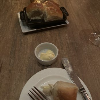 Mooo.... Beacon Hill - Parker rolls - A piece of bread with butter on a white plate, a small bowl of butter, and a glass of wine on a wooden table, with a candle in the background. - Steakhouses Near Me - Boston, Massachusetts