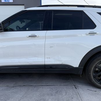 Body Master - Car - A white SUV is parked on a concrete surface, with visible scratches and dents on the driver's side door. The vehicle has tinted windows and black wheels, with a building and a garage door in the background. - Body Shops Near Me - Albuquerque, New Mexico