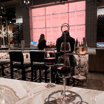 Apericena Wine Bar - Interior bar area - A bar interior features a marble table with three wine glasses, two filled with red wine and one with white wine, placed on a metal wine glass holder. Behind the bar, a pink-lit wall displays hanging wine glasses and bottles, with two staff members preparing drinks. The setting includes black chairs with striped cushions, decorative hanging lanterns, and wine racks on the side. The atmosphere suggests a sophisticated wine bar environment. - Wine Bars Near Me - Upper Saint Clair, Pennsylvania