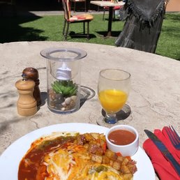 Huevos Rancheros - A breakfast plate featuring huevos rancheros topped with melted cheese, accompanied by salsa and crispy diced potatoes, is placed on a stone table outdoors. The table also holds a glass of orange juice, a small candle holder with a succulent plant, and salt and pepper shakers. In the background, there are tables and chairs set in a garden area, with a statue of a woman and a shaded patio visible.