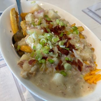 The Parrot - Clam Chowder Fries - A white oval dish contains clam chowder fries topped with chopped green onions and bacon bits. The fries are covered with a creamy clam chowder sauce, and a metal spoon is placed inside the dish. The dish is on a table with a menu and glasses in the background, indicating a restaurant setting. - Seafood Near Me - Hull, Massachusetts