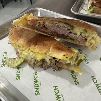 Adriano's Sunset - The Cuban - A toasted sandwich with sliced meat, melted cheese, and lettuce is placed on a metal tray lined with branded paper that reads 'SANDWICH MIAMI.' The sandwich is cut in half, revealing the ingredients inside. The background shows a dining area with chairs and people, indicating a casual restaurant setting. - Sandwiches Near Me - Miami, Florida
