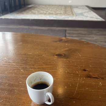 Black Dog Vinyl Cafe - Espresso. Very dark and bold intense flavor. - A small white ceramic coffee cup containing black coffee is placed on a wooden table. The cup has coffee stains on its inner rim, indicating it has been used. The table surface shows visible wood grain and some scratches. In the background, there is a carpeted area with a patterned rug and a few chairs, suggesting an indoor setting such as a cafe or home. - Coffee & Tea Near Me - Plainfield, Illinois