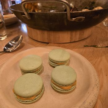 Mariscos San Pedro - Macarons! - Four green macarons with filling are placed on a round, textured wooden plate on a restaurant table. The table has various items including a spoon, glasses, and a large black skillet in the background. The setting appears to be a dining environment with other diners visible in the background. - Mexican Near Me - Chicago, Illinois