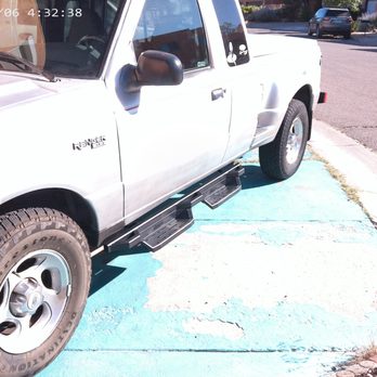 Auto Depot - A white Ford Ranger pickup truck is parked on a light blue painted parking space in an outdoor lot. The truck has black side mirrors and black running boards. The image shows part of the front and side of the vehicle, with a background of other parked cars and a street with buildings and vegetation. The photo was taken during daylight hours, with shadows indicating sunlight. - Auto Detailing Near Me - Albuquerque, New Mexico