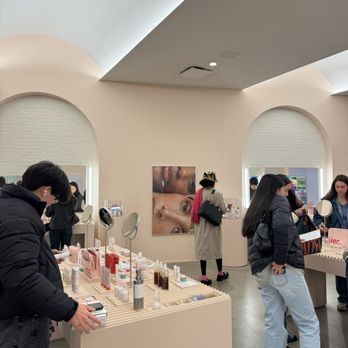 Glossier Chicago - A retail space with customers examining skincare, makeup, and fragrance products on display tables, featuring mirrors and modern decor. - Cosmetics & Beauty Supply Near Me - Chicago, Illinois