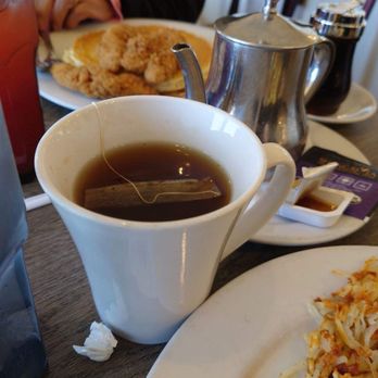 Buttered Toast Restaurant - The image shows a table setting at a restaurant with a white ceramic cup containing tea with a tea bag inside, a plate of coleslaw, and a glass of water. A metal teapot and a small dish of honey or syrup are also visible on the table. In the background, there is a plate of fried food, possibly chicken or fish, and some condiments. The setting appears to be casual dining, with a focus on breakfast or brunch items. - Breakfast & Brunch Near Me - Algonquin, Illinois