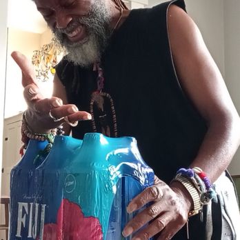 Walmart Supercenter - My Favorite Place for keeping a good balance with water that's "highly beneficial" for my Blood-type!! - A man with a beard and dark skin, wearing a sleeveless black shirt, is smiling and pointing at a pack of bottled water on a wooden table. The pack is blue and pink, and the man is wearing multiple bracelets on his wrist. The background shows a light-colored wall and a doorway with a window. - Grocery Near Me - Dixon, Illinois